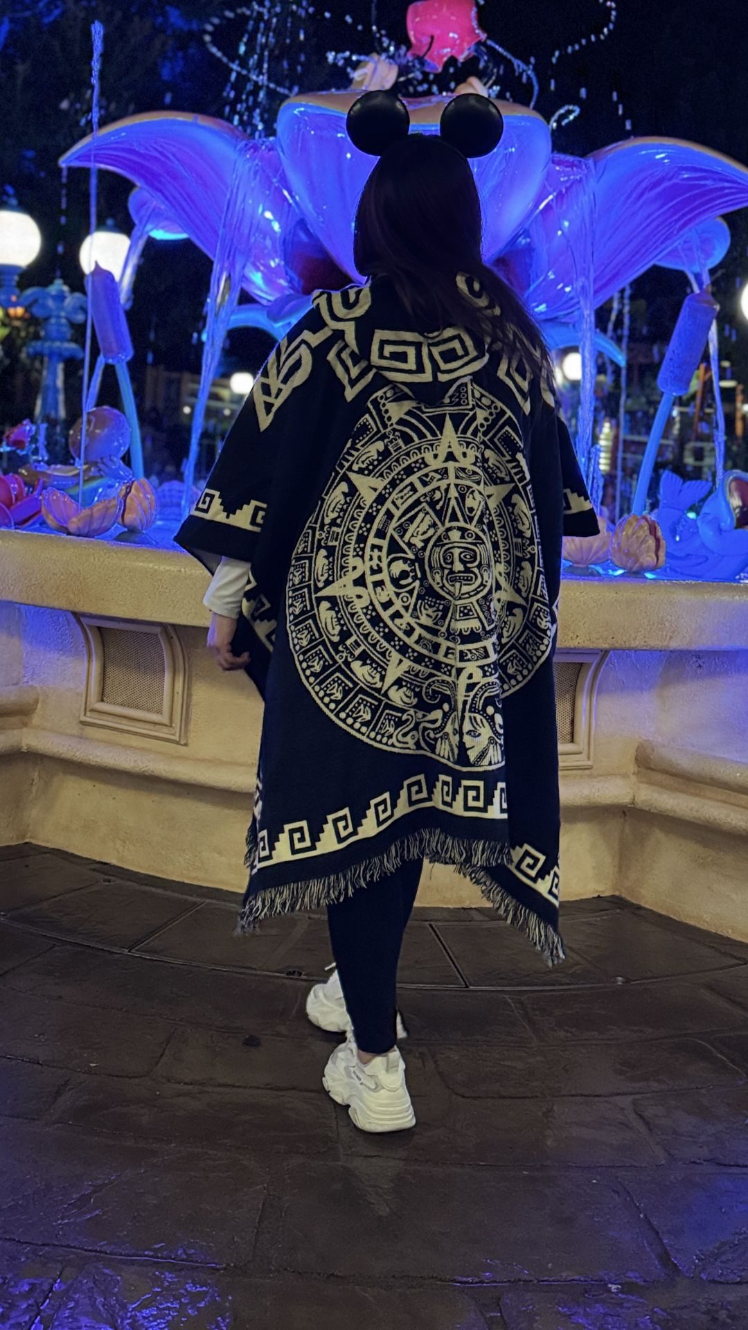 Crystal in an Aztec calendar poncho and mouse ears at a blue-lit fountain after dark