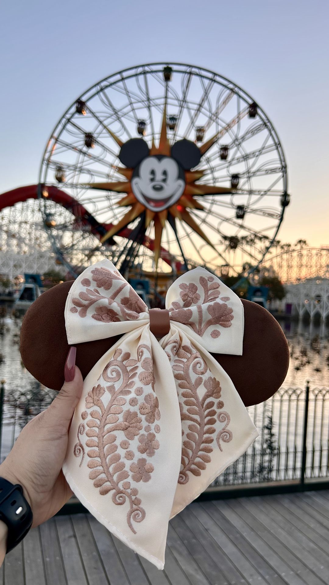 Brown Minnie ears with a cream otomí-embroidered bow held up against the Pixar Pier wheel at sunset