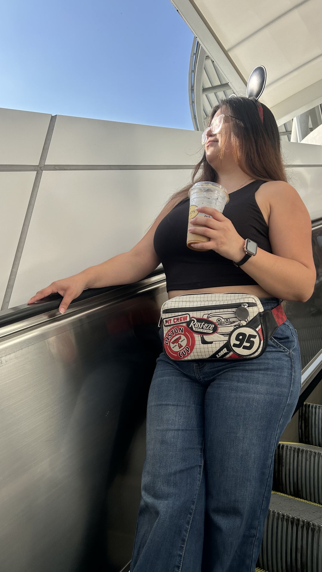 Crystal on an escalator with a Piston Cup belt bag, mouse ears and an iced coffee