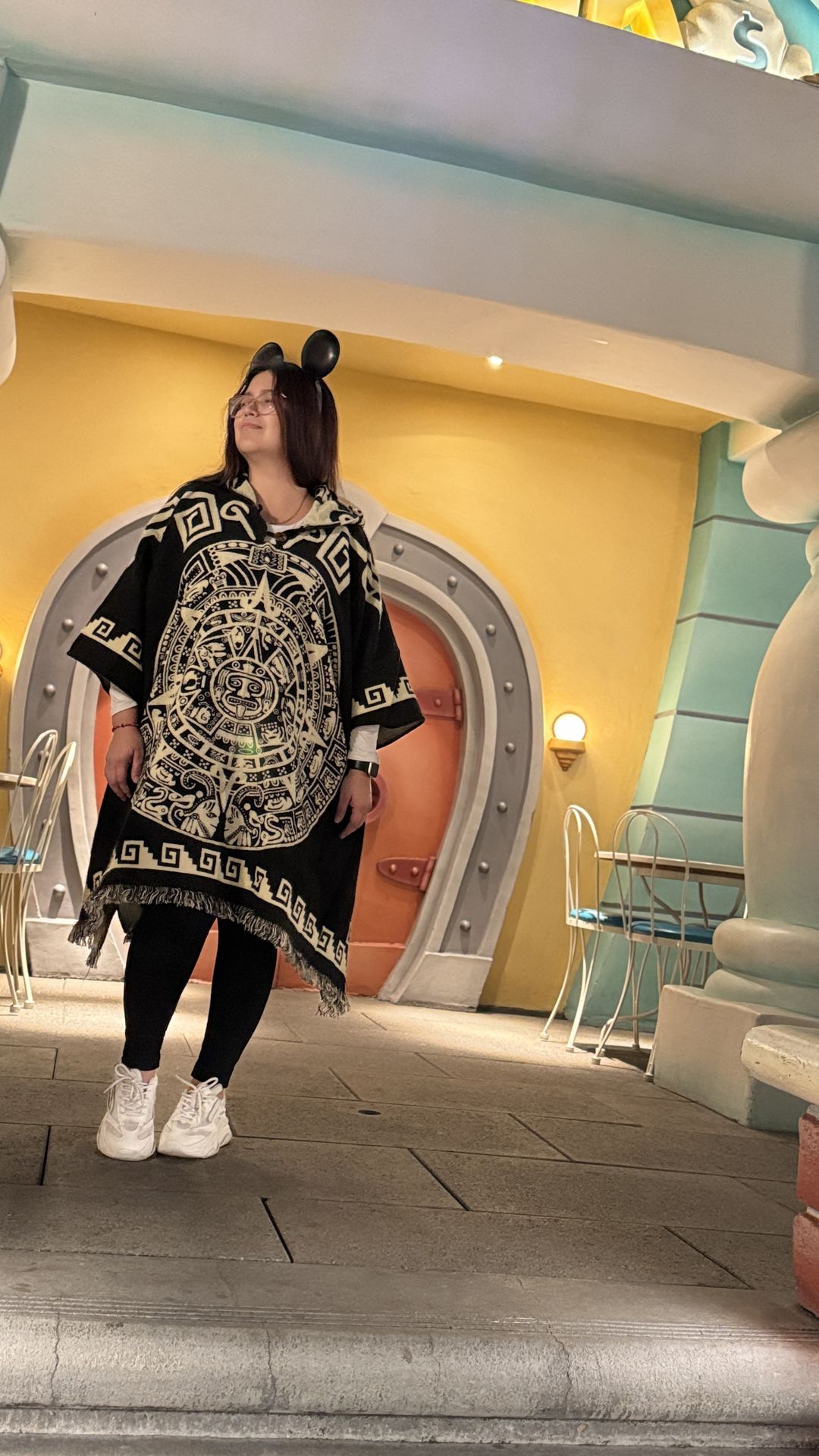 Crystal in an Aztec calendar poncho and mouse ears outside a pastel Toontown storefront